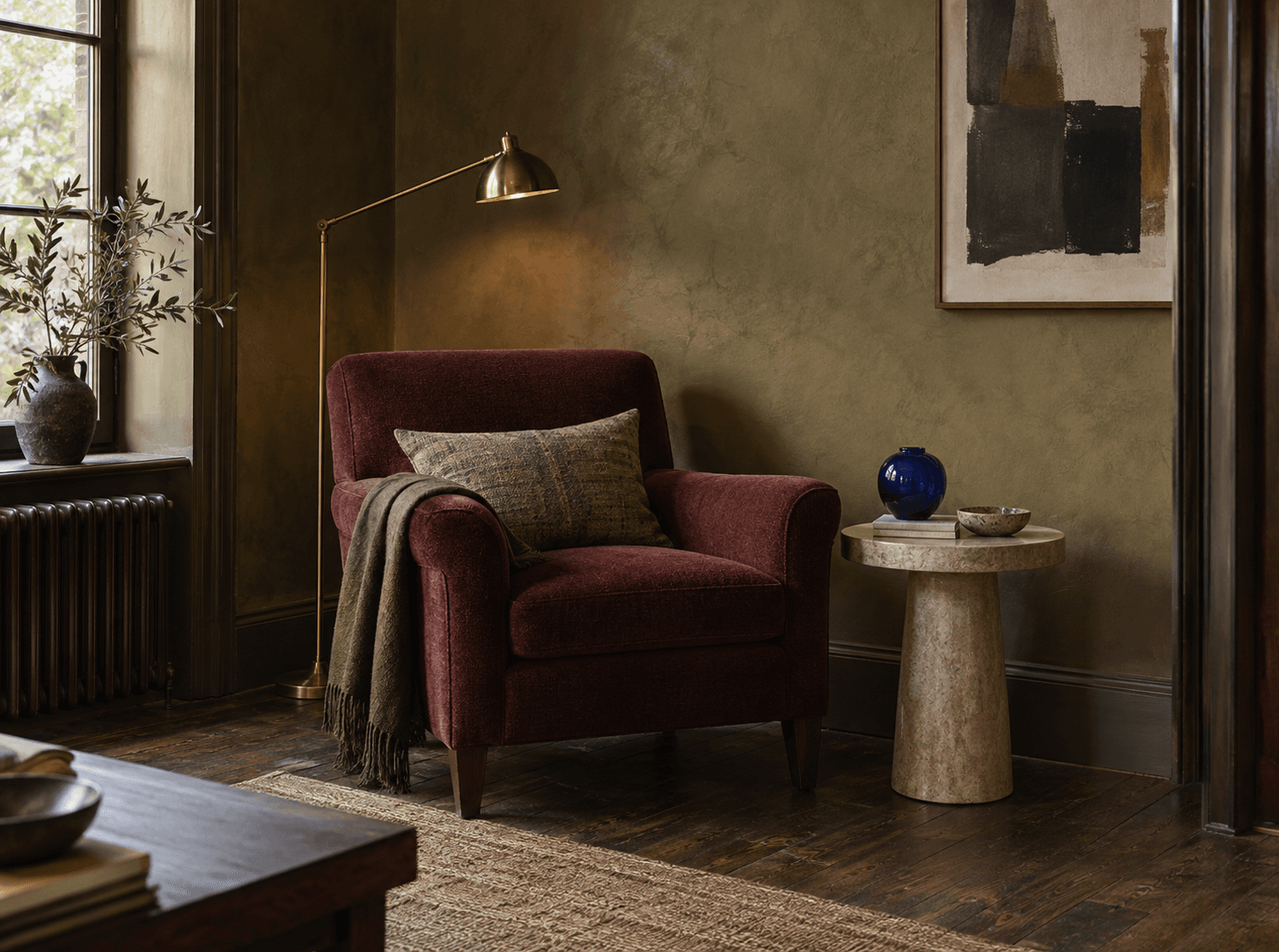 Conceptual reading corner with an olive plaster wall, oxblood chair, brass lamp, stone table, and cobalt glass vessel.