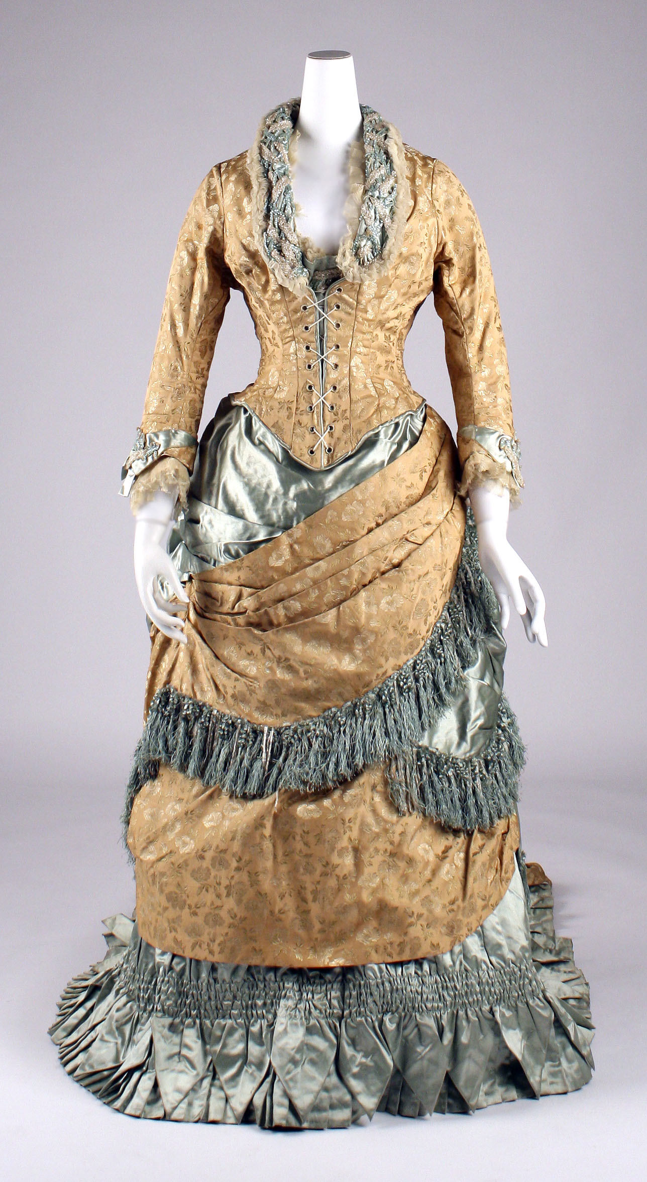An American dress from 1880 has a fitted laced bodice, layered gold and pale blue fabric, fringe and a pleated hem.